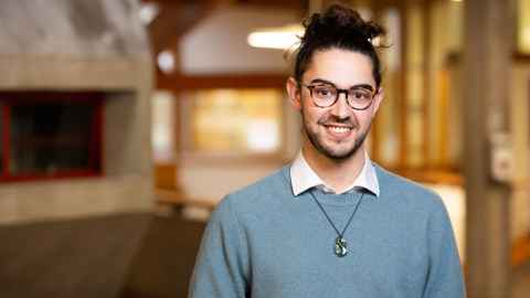 Music and Global Studies student standing in building foyer wearing a grey sweatshirt and pounamu smiling at camera.